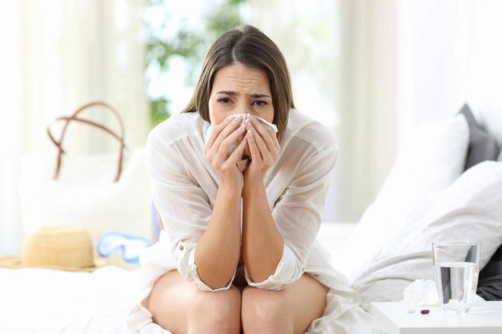 Front view portrait of an ill allergic woman looking at camera sitting on the bed of an hotel room on summer vacations